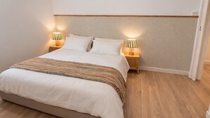 A neatly made bed with a white comforter and a striped blanket, two bedside lamps, and a wooden floor.