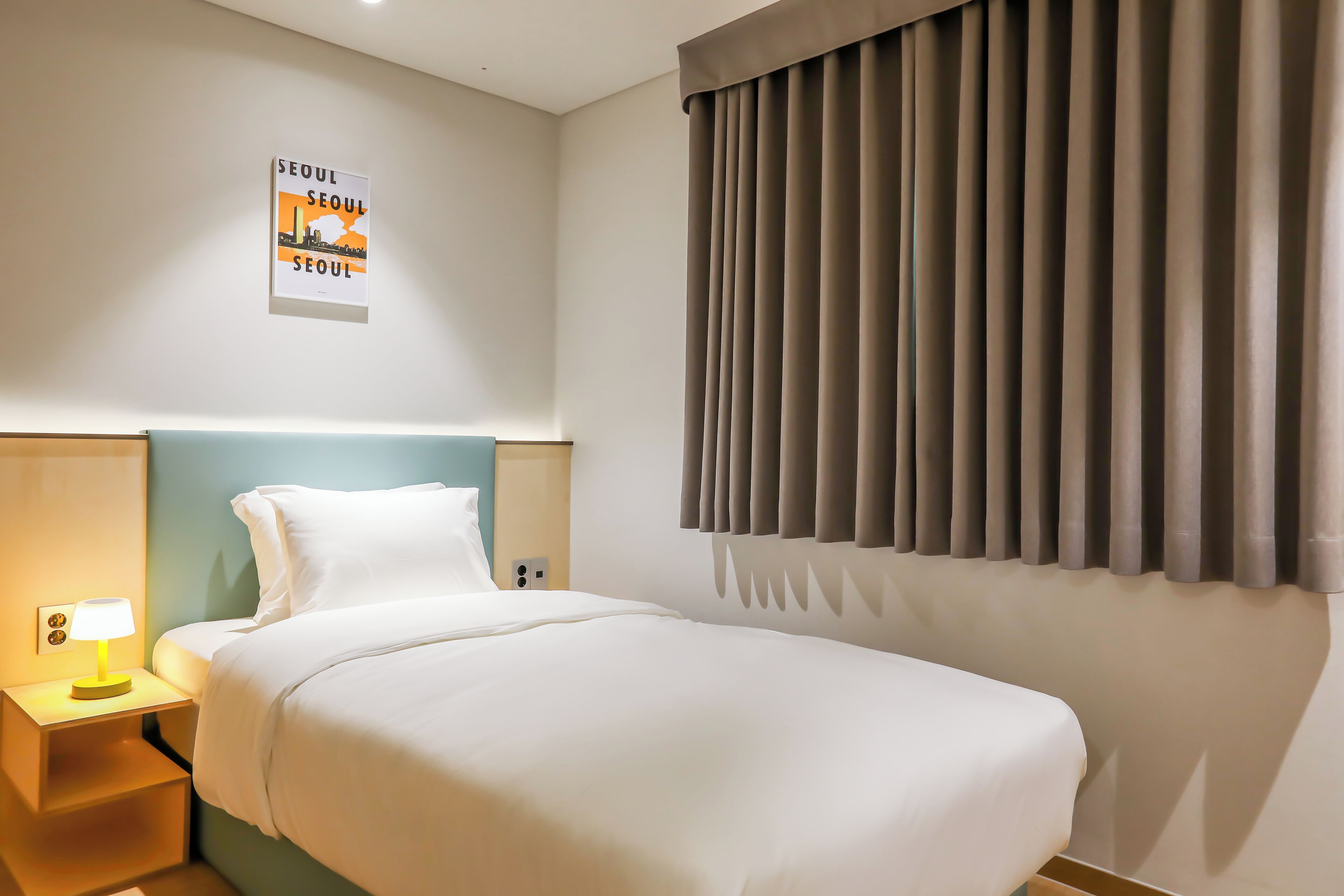 Habitación de hotel con cama, mesita de noche con lámpara, cartel en la pared y ventana con cortinas.
