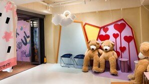 A room with a large teddy bear, a smaller teddy bear, and a third teddy bear on a table. There is a chair and a small stool.