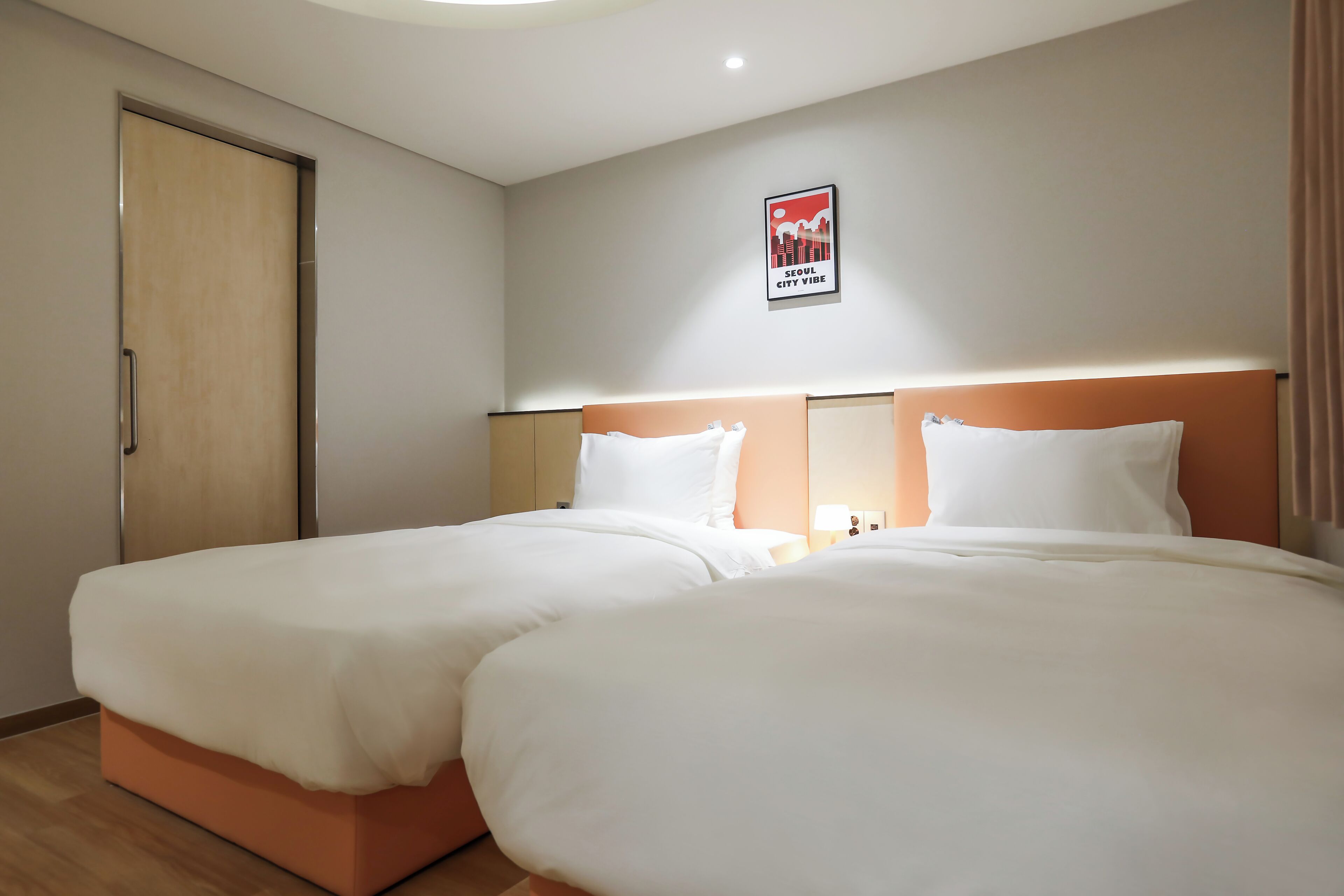 Une chambre d’hôtel avec deux lits, une porte, une affiche au mur et une table de chevet avec une lampe.