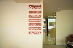 Una pared con varios letreros que indican normas como vigilancia por circuito cerrado, prohibido fumar, no se permiten visitantes, comprobante de identificación obligatorio, no se permite comida de fuera, prohibido porte de armas y municiones, y horarios de registro de entrada y salida.