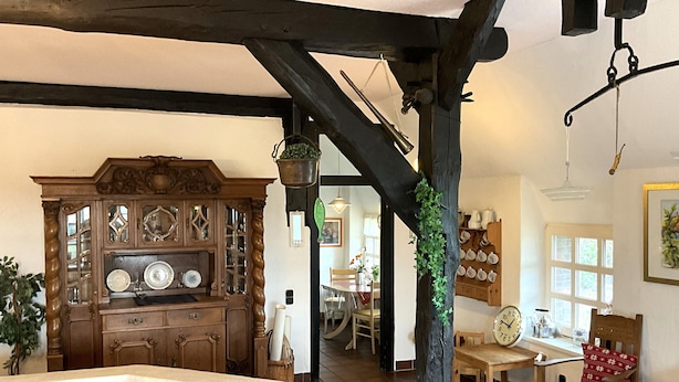 Ein traditionelles Interieur mit freiliegenden Holzbalken, einem Holzschrank mit Glastüren, einer Essecke mit Tisch und Stühlen sowie einer hängenden Pflanze.