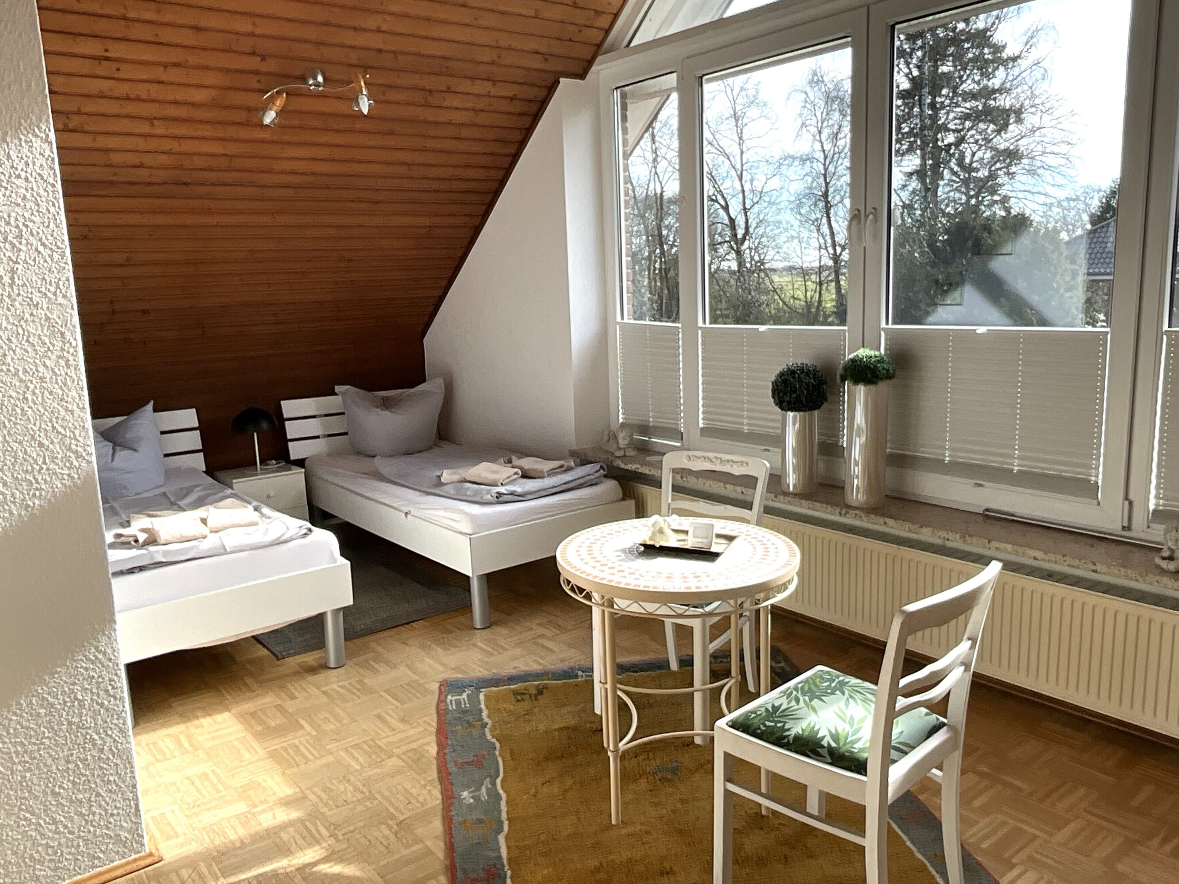 A bedroom with a sloped ceiling, a bed, a chair, a table, and a window with blinds.