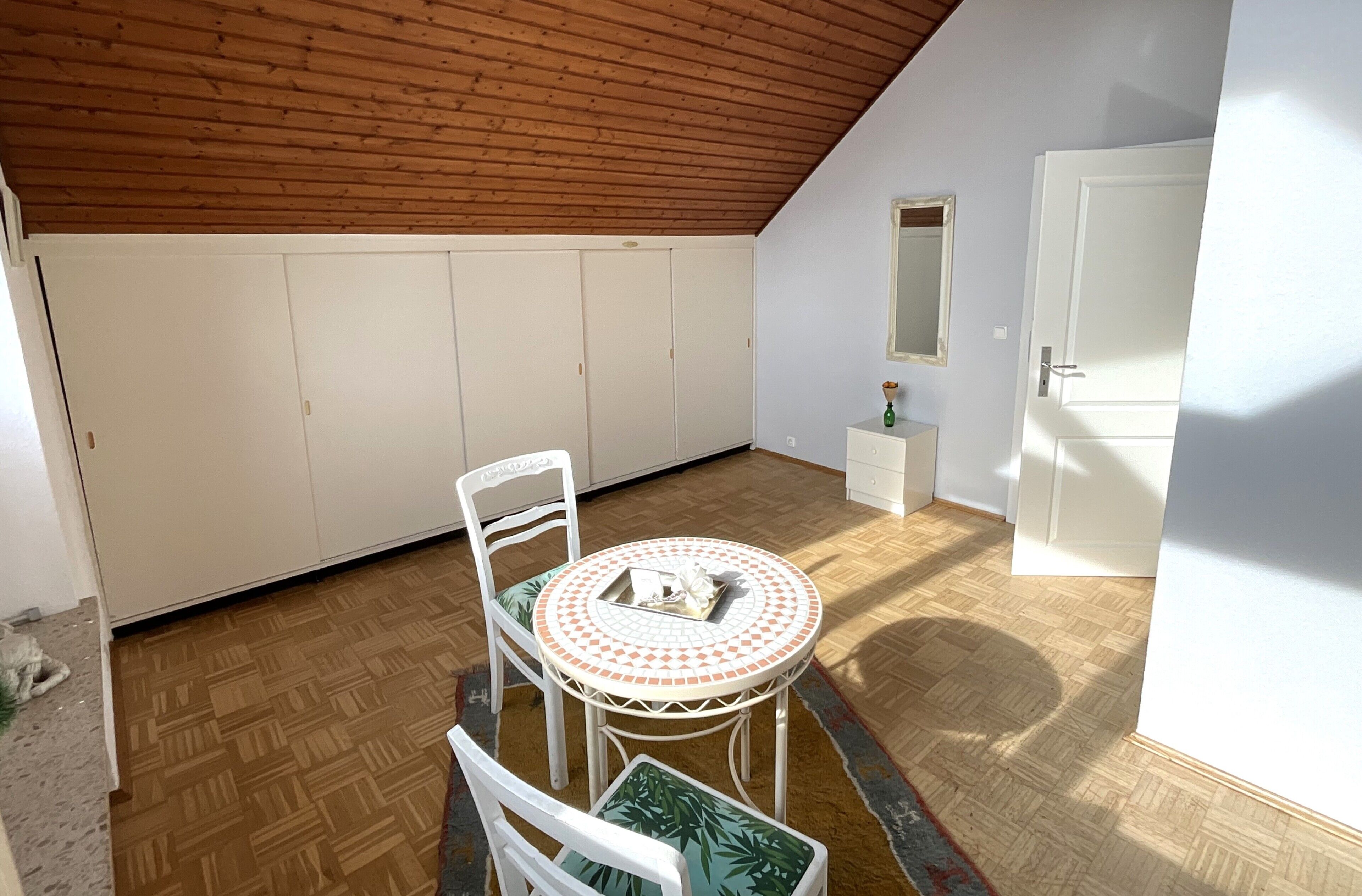 A room with a wooden ceiling, a white wardrobe, a round table with chairs, a small white cabinet, and a door.