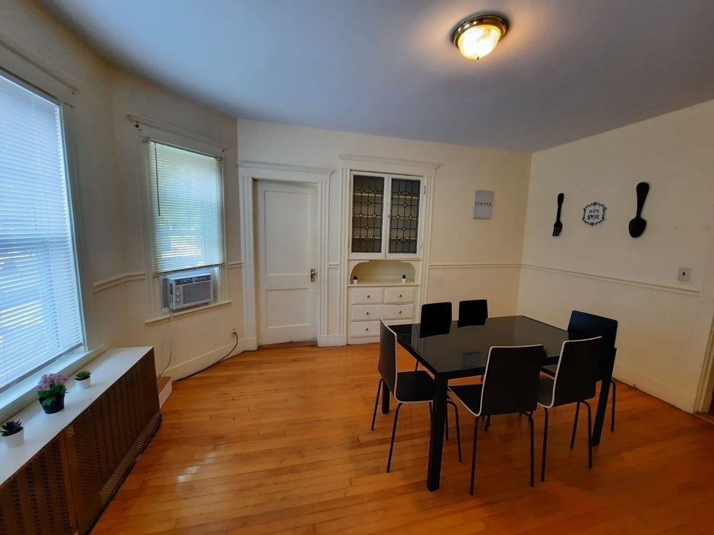 A dining area with a table and chairs, a radiator, a window with blinds, a door, and a wall-mounted light fixture.