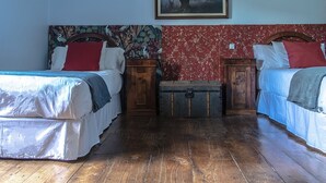 Ein Zimmer mit zwei Betten, Holzboden, einem blumigen Kopfteil, einer rot gemusterten Wand und einem gerahmten Bild.