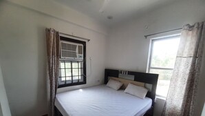 A single bed with a headboard, a window with a curtain, and a wall-mounted air conditioning unit. - Hotel Aditya Bikaner (Bikaner)