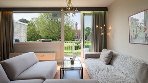 En moderne stue med sofa, sofabord og en balkon med udsigt over grøn natur og bygninger.