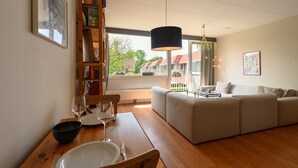 En stue med sofa, spisebord med stole, bogreol og et stort vindue med udsigt til huse.