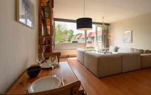 Ein Wohnzimmer mit einer Couch, einem Esstisch mit Stühlen, einem Bücherregal und einem großen Fenster mit Blick auf Häuser.