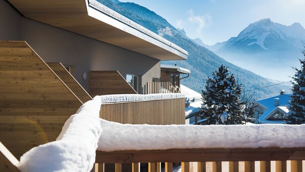 A wooden deck with snow, a mountain view, and a building with a sloped roof.