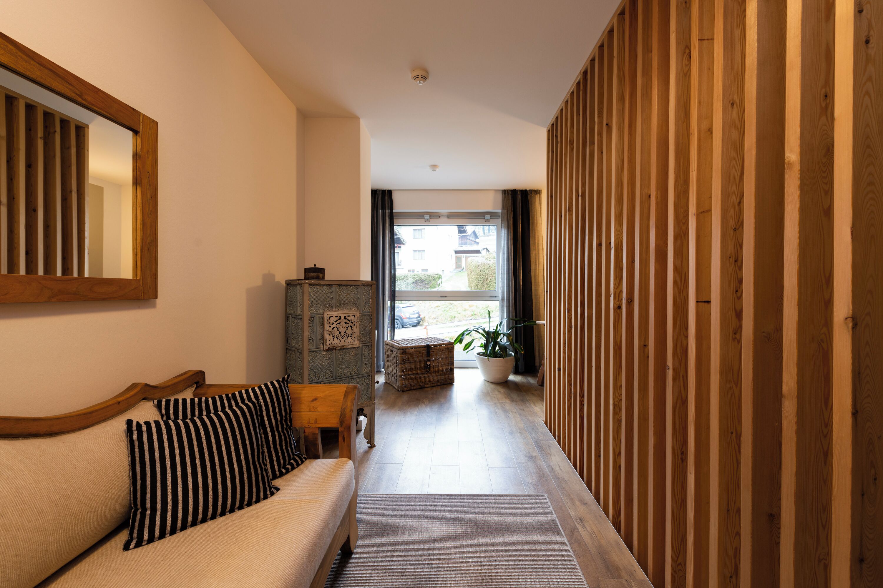 A hallway with a wooden floor, a sofa with striped pillows, a wooden wall panel, and a large window.