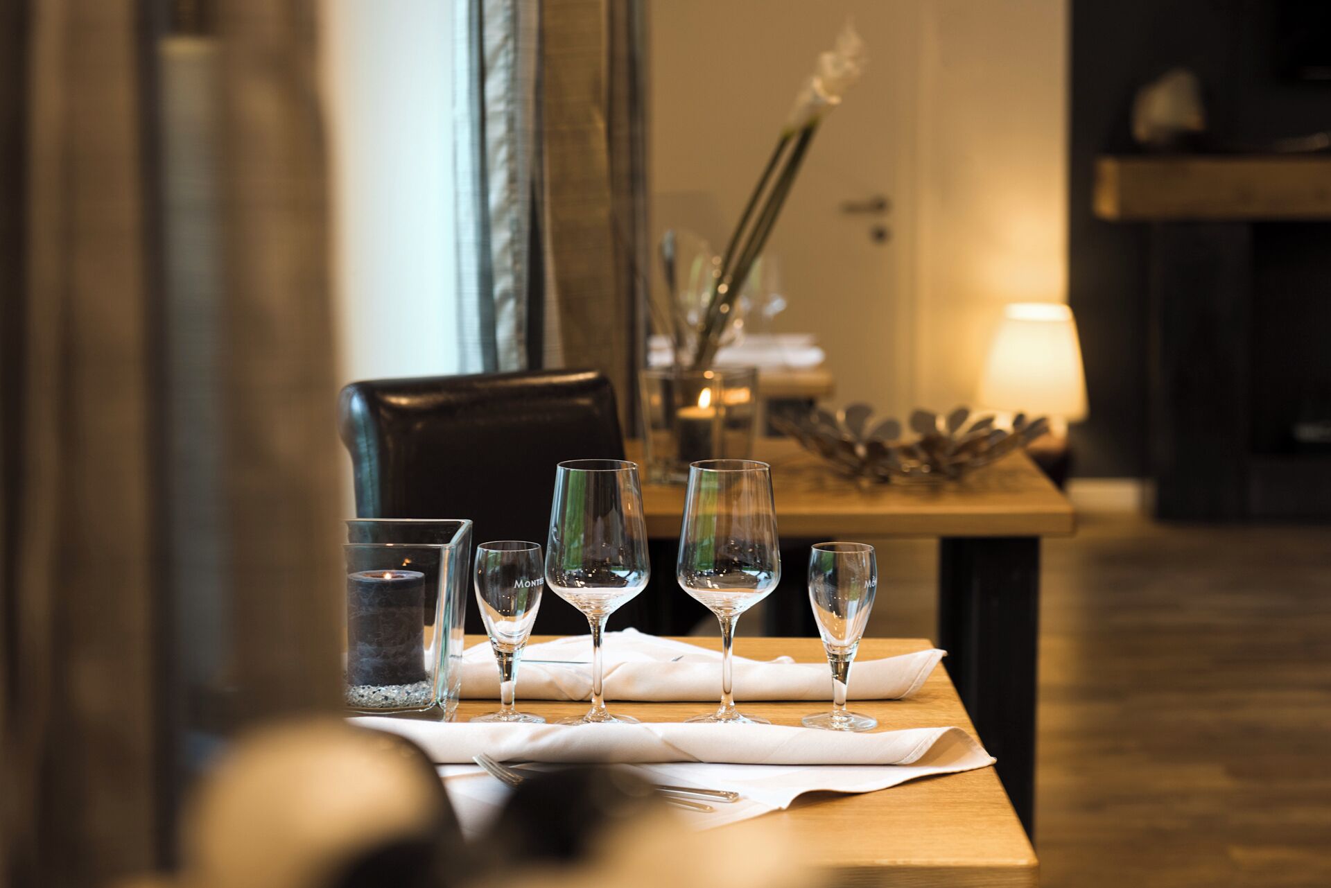 Een eettafel met verschillende wijnglazen en een kaars, geplaatst in een zwak verlichte kamer met een spiegel en een lamp op de achtergrond.