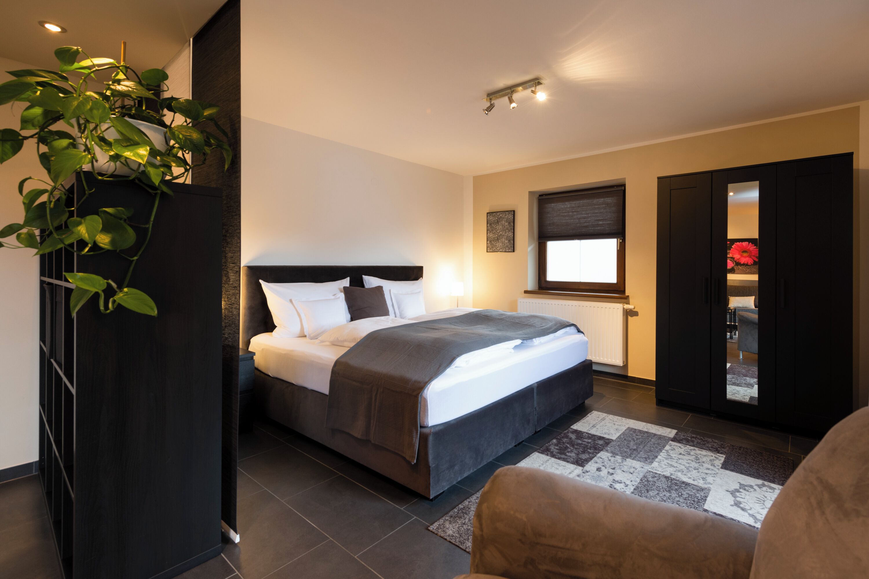 A modern bedroom with a bed, a potted plant, a window, and a door leading to another room.