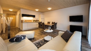 A modern living room with a sofa, a coffee table, a dining table with chairs, a kitchen area with a sink and cabinets, and a flat-screen TV mounted on the wall.