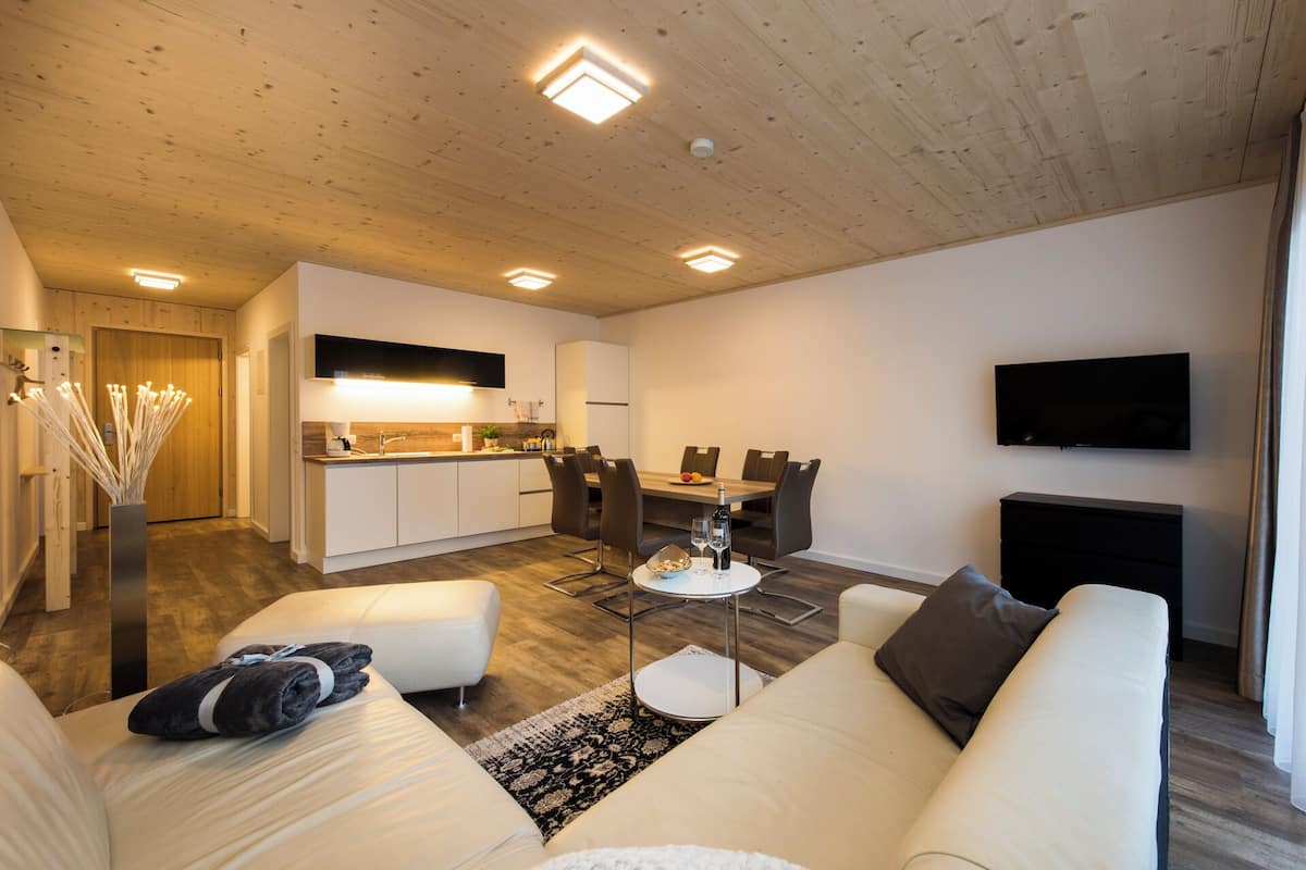 Una sala de estar moderna con un sofá, una mesa de centro, una mesa de comedor con sillas, un área de cocina con un fregadero y armarios, y un televisor de pantalla plana montado en la pared.