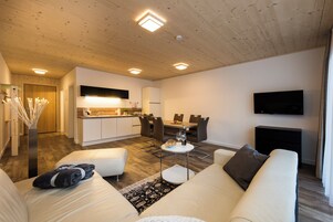 Ein modernes Wohnzimmer mit Sofa, Couchtisch, Esstisch mit Stühlen, Küchenzeile mit Spüle und Schränken sowie einem wandmontierten Flachbildfernseher.