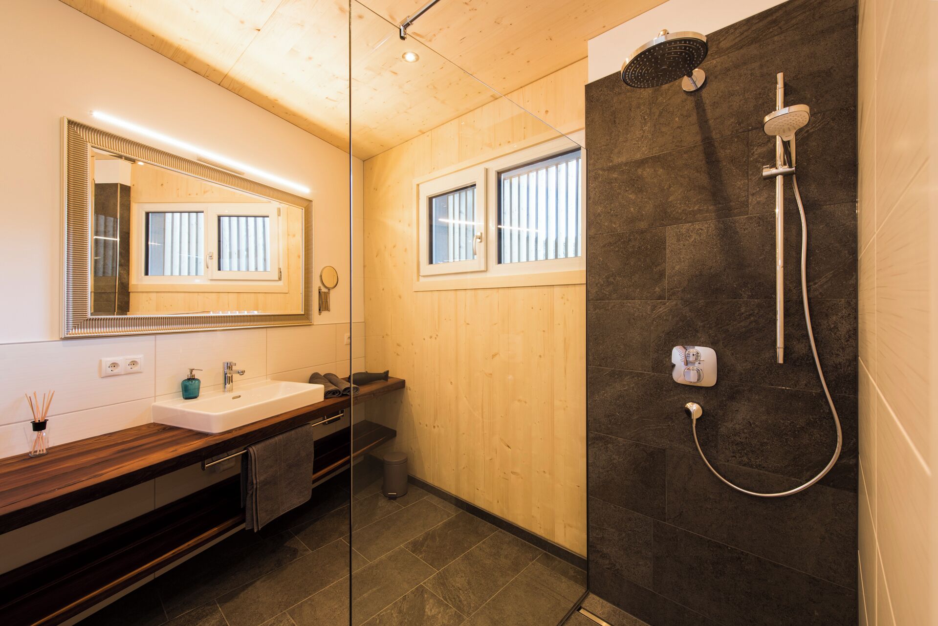 A modern bathroom with a wooden vanity, a large mirror, a shower with a control panel, and a window.