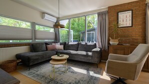 A modern living room with a sectional sofa, a round coffee table, a chair, and a large window with a view of greenery. - Holiday home Salvatorhoeve 120 nature house at Salvatorhoeve (Ouwerkerk)