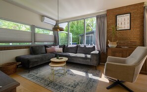 A modern living room with a sectional sofa, a round coffee table, a chair, and a large window with a view of greenery. - Holiday home Salvatorhoeve 120 nature house at Salvatorhoeve (Ouwerkerk)