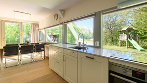 A modern kitchen with a large window, a dining area with chairs, and a view of a playground. - Holiday home Salvatorhoeve 120 nature house at Salvatorhoeve (Ouwerkerk)