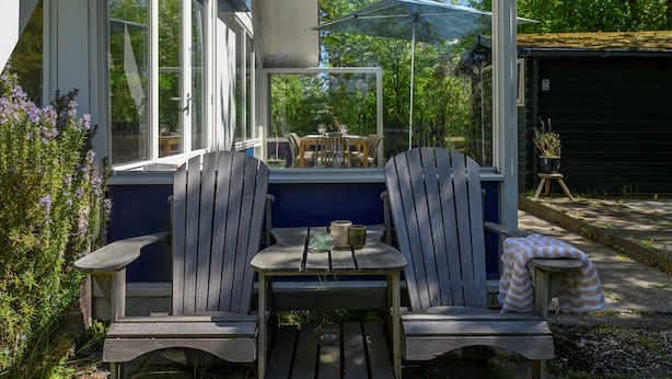 A wooden deck with two Adirondack chairs, a small table with a cup and saucer, and a covered patio area. - Holiday home Salvatorhoeve 120 nature house at Salvatorhoeve (Ouwerkerk)