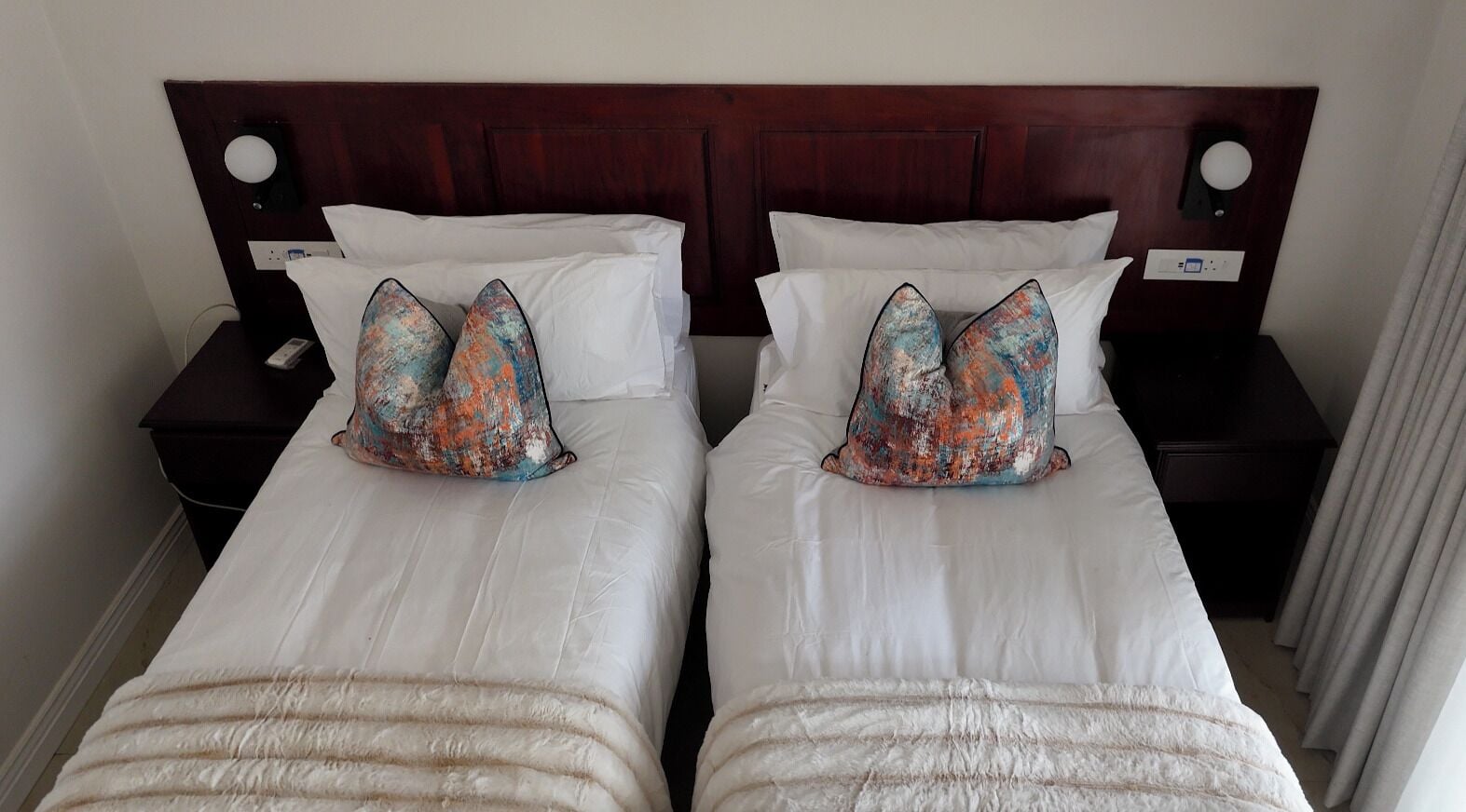 Un lit double avec du linge de lit blanc et deux coussins décoratifs, une tête de lit en bois et une table de chevet avec une lampe.