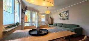 A modern living room with a wooden dining table, a green sofa, and a large window with blinds. - Vacation apartment i37 - Spacious vacation apartment with balcony, elevator and modern (Graal-Müritz)
