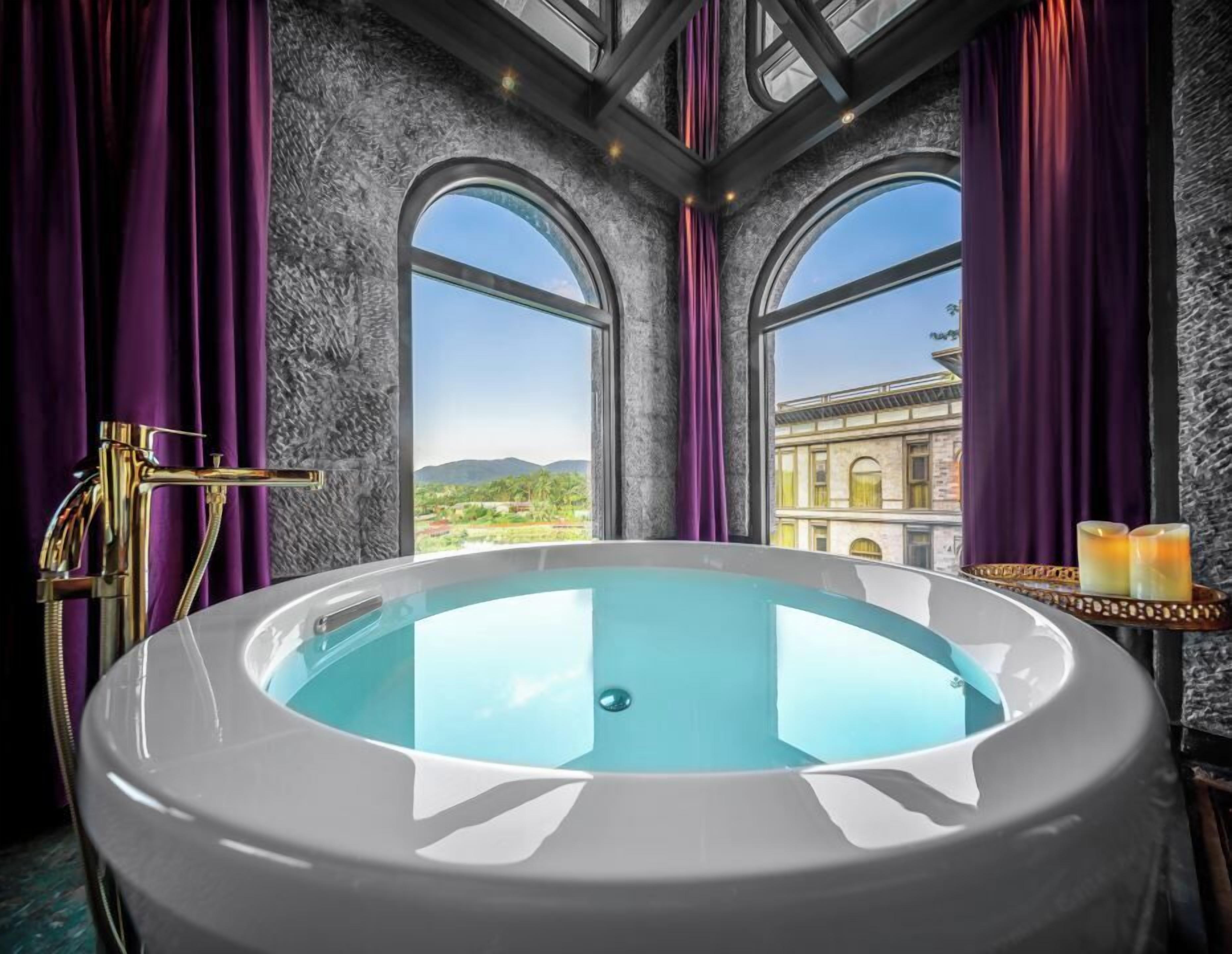 Una bañera grande con vista a un edificio a través de ventanales arqueados, cortinas moradas y un grifo dorado.