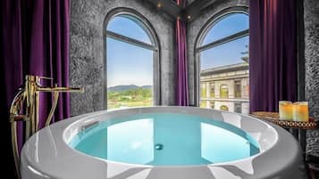 Una bañera grande con vista a un edificio a través de ventanales arqueados, cortinas moradas y un grifo dorado.