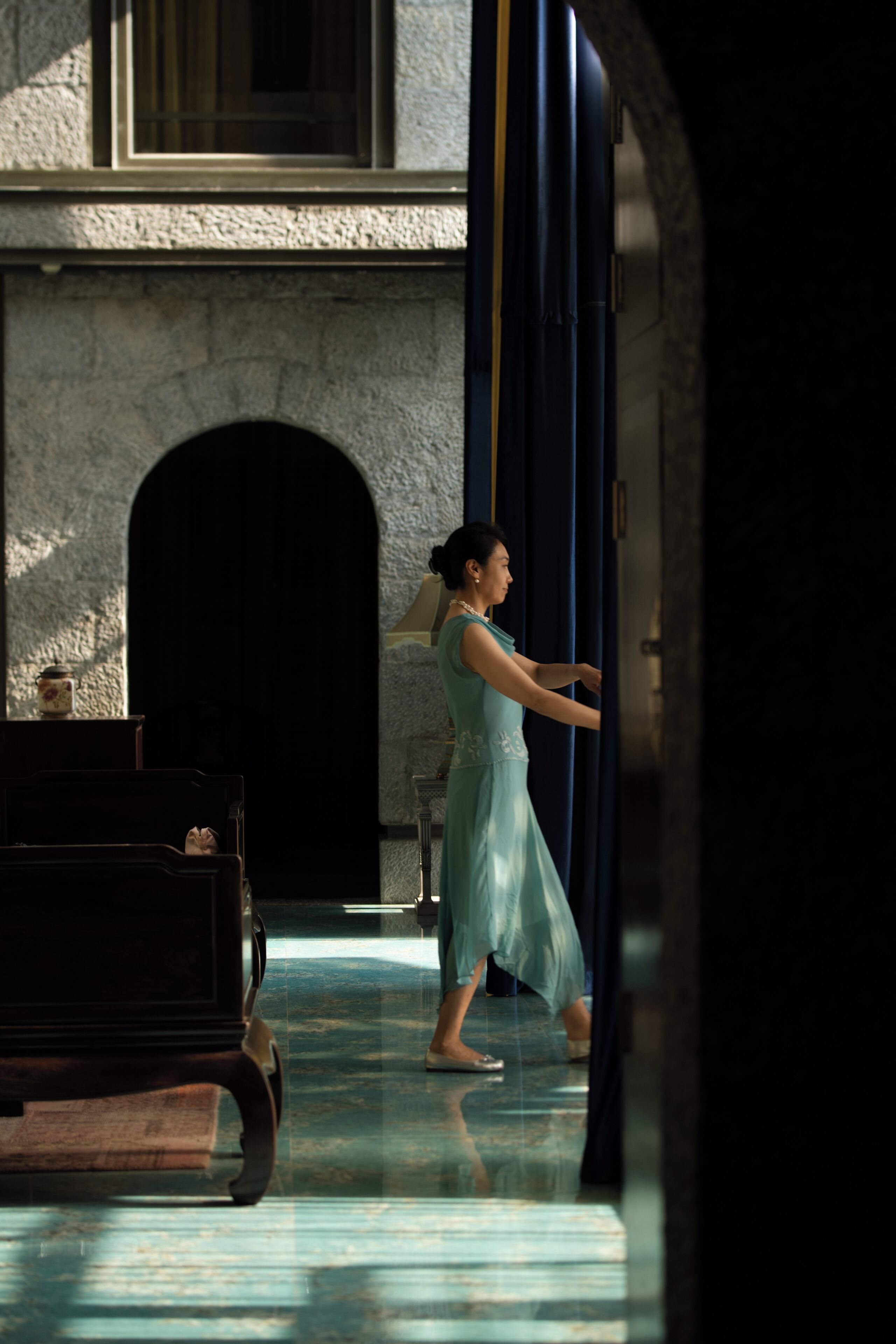 A woman in a dress standing in a doorway, with a stone building and a bench in the background.