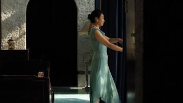 A woman in a dress standing in a doorway, with a stone building and a bench in the background.
