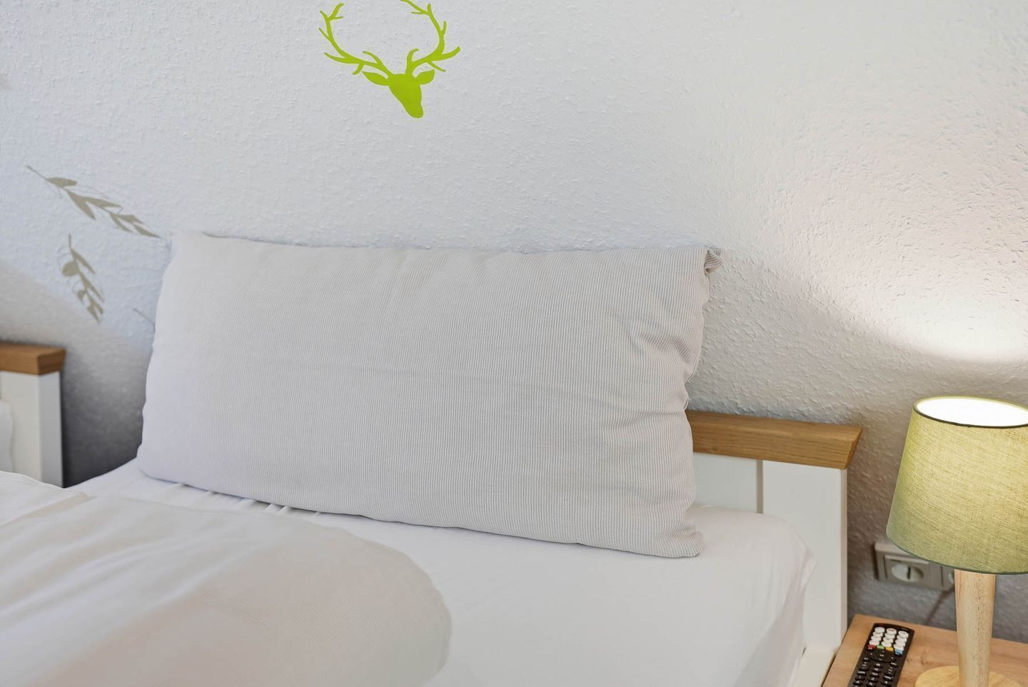 Un lit avec un oreiller à motifs, un autocollant représentant un cerf accroché au mur, une lampe et une télécommande posées sur une table de chevet.