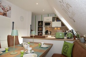 A cozy kitchen with a dining area, featuring a green kettle, a clock, and a decorative deer figurine. - Platzhirsch - New Platzhirsch vacation apartment in Greetsiel for relaxed (Krummhörn)