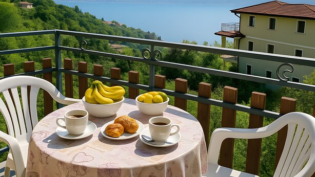 A balcony with a table set for two, featuring a floral tablecloth, two white chairs, a bowl of bananas, a bowl of lemons, a croissant, and two cups of coffee. - Cozy 90 sqm apartment in Tignale with pool, garden, terrace, parking and Wi-Fi (Aer)