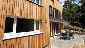 Ein modernes Holzgebäude mit großen Fenstern, einem Balkon und einer Terrassenfläche mit Sitzgelegenheiten.