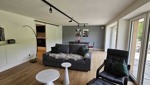 Ein modernes Wohnzimmer mit grauem Sofa, einem kleinen runden Tisch und einem großen Fenster mit Blick ins Freie.