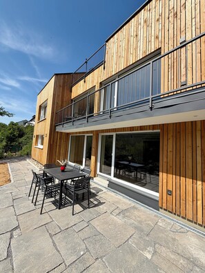 Un bâtiment moderne doté d’une façade en bois, de grandes fenêtres et d’un patio aménagé avec des sièges extérieurs.