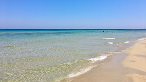 Ein Strand mit klarem Wasser, sandigem Ufer und einem fernen Segelboot.
