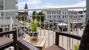 A balcony with wicker furniture, a potted plant, and a view of a town square with buildings and outdoor seating. - Syltliebe - Modern and central app. for 4 persons with balcony, car (Sylt)