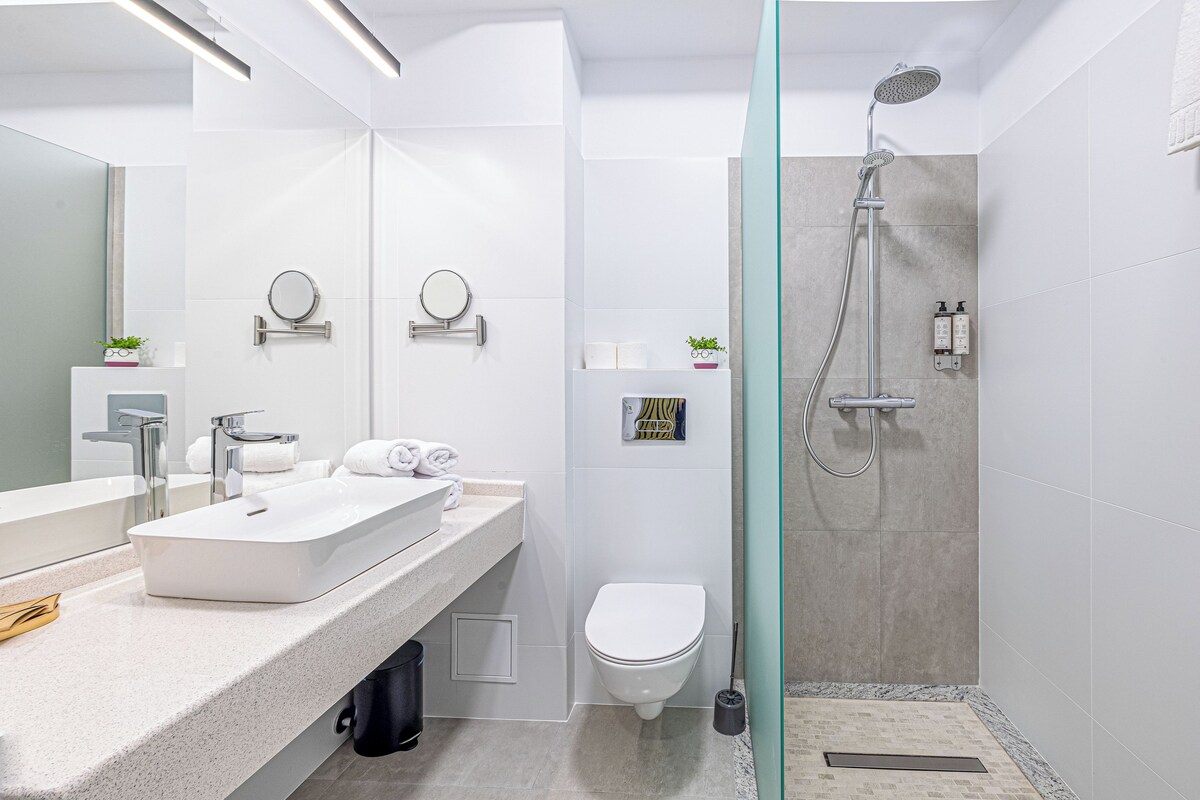 A modern bathroom with a large mirror, a white sink, a toilet, and a shower area.