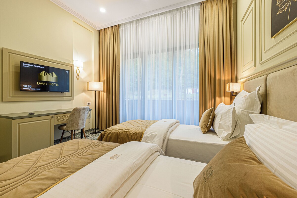 Une chambre d’hôtel avec deux lits, un bureau, une chaise, un téléviseur affichant le nom de l’hôtel et une grande fenêtre avec des rideaux.