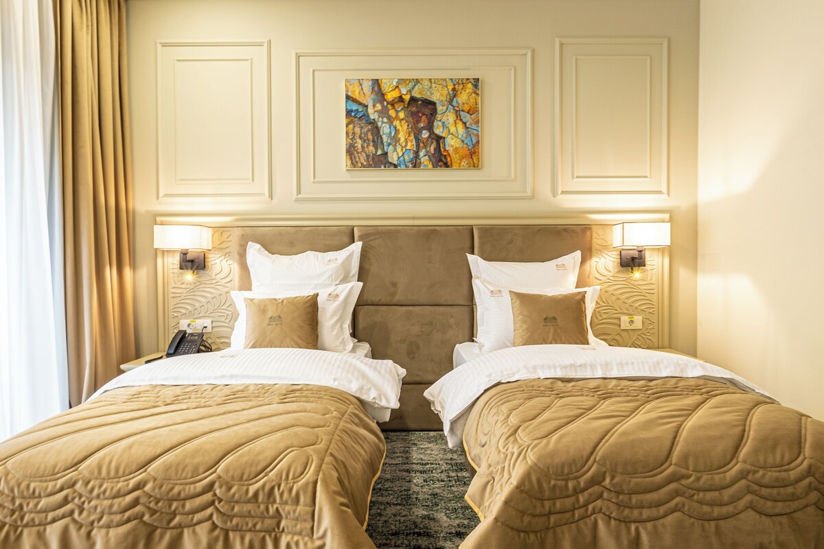 A hotel room with two beds, beige bedding, a painting on the wall, and bedside lamps.