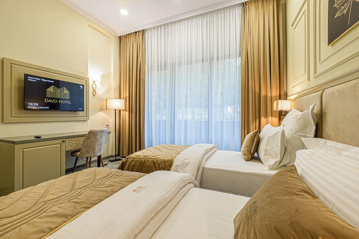 Camera d'albergo con due letti, una scrivania, una sedia, un televisore che mostra il nome dell'hotel e un'ampia finestra con tende.