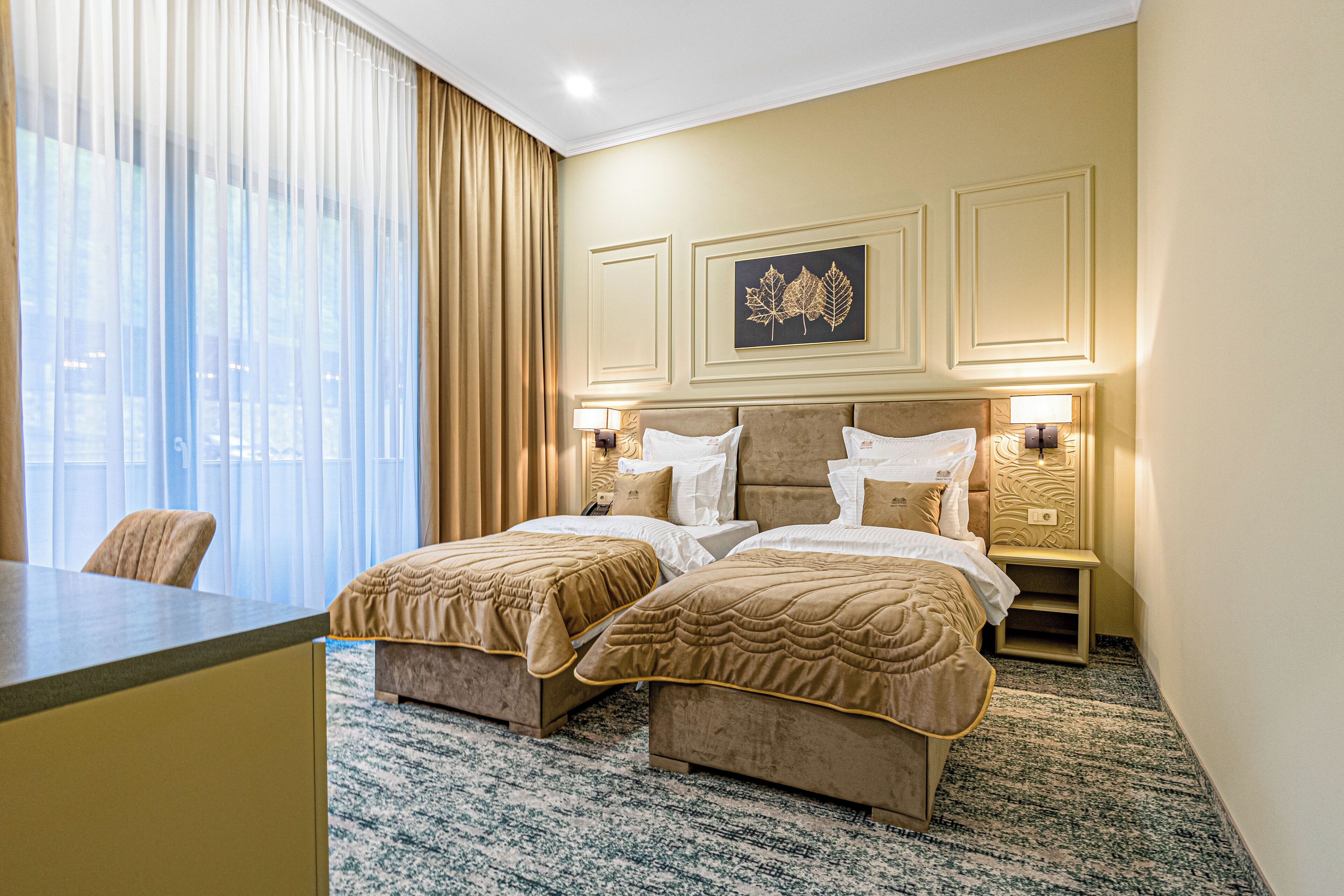 A hotel room with two beds, a desk, a chair, a large window with curtains, and a wall-mounted artwork.