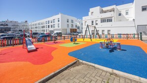 A playground with colorful play areas and a red slide, surrounded by a fence and parked cars. - Holiday Home 'Apartamento Bahía Serena Conil' with Wi-Fi (Conil de la Frontera)