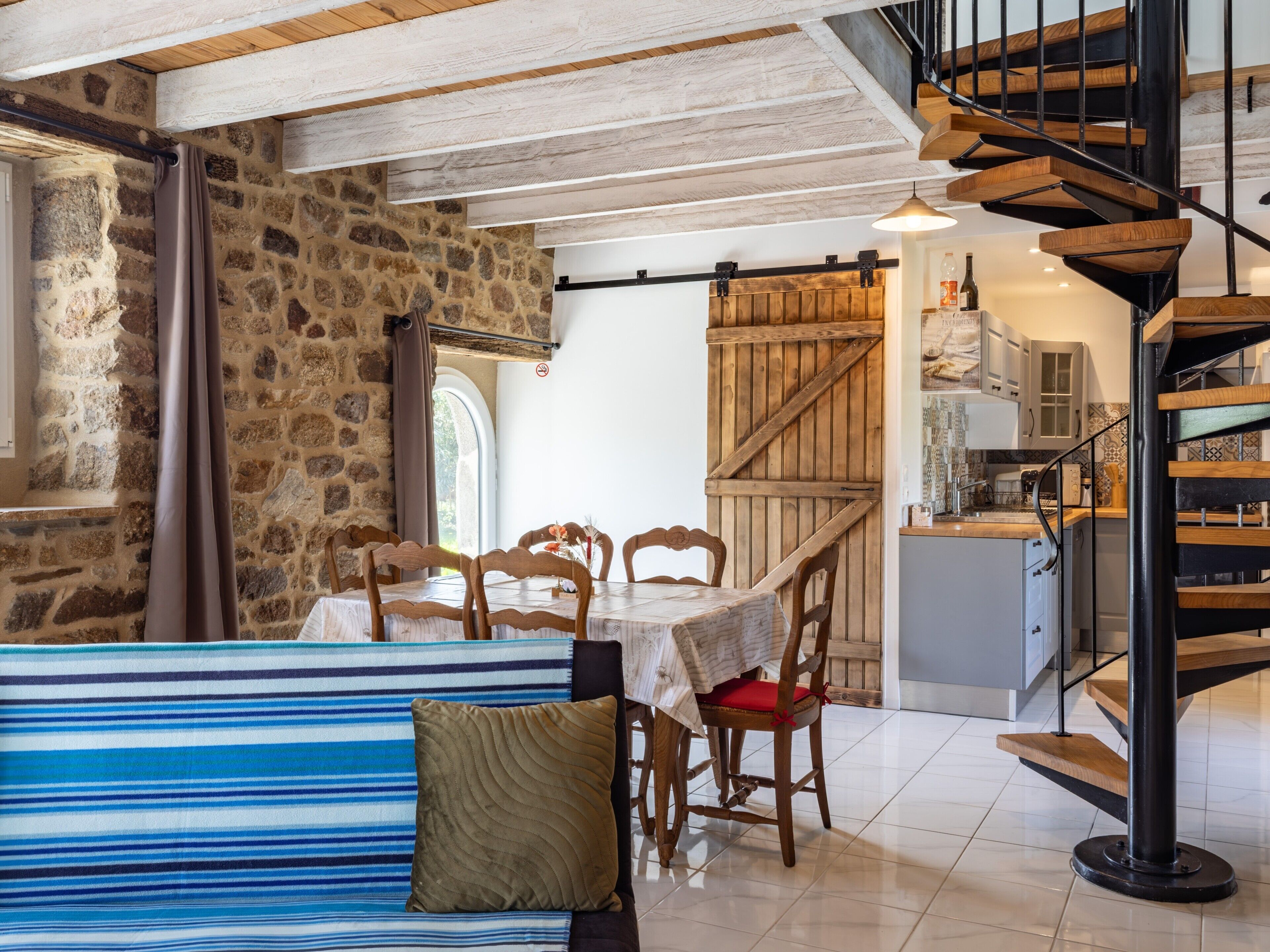 Ein Essbereich mit einem Holztisch, Stühlen und einer gestreiften Couch. Eine Treppe mit Holzwand verläuft zu einer erhöhten Ebene.
