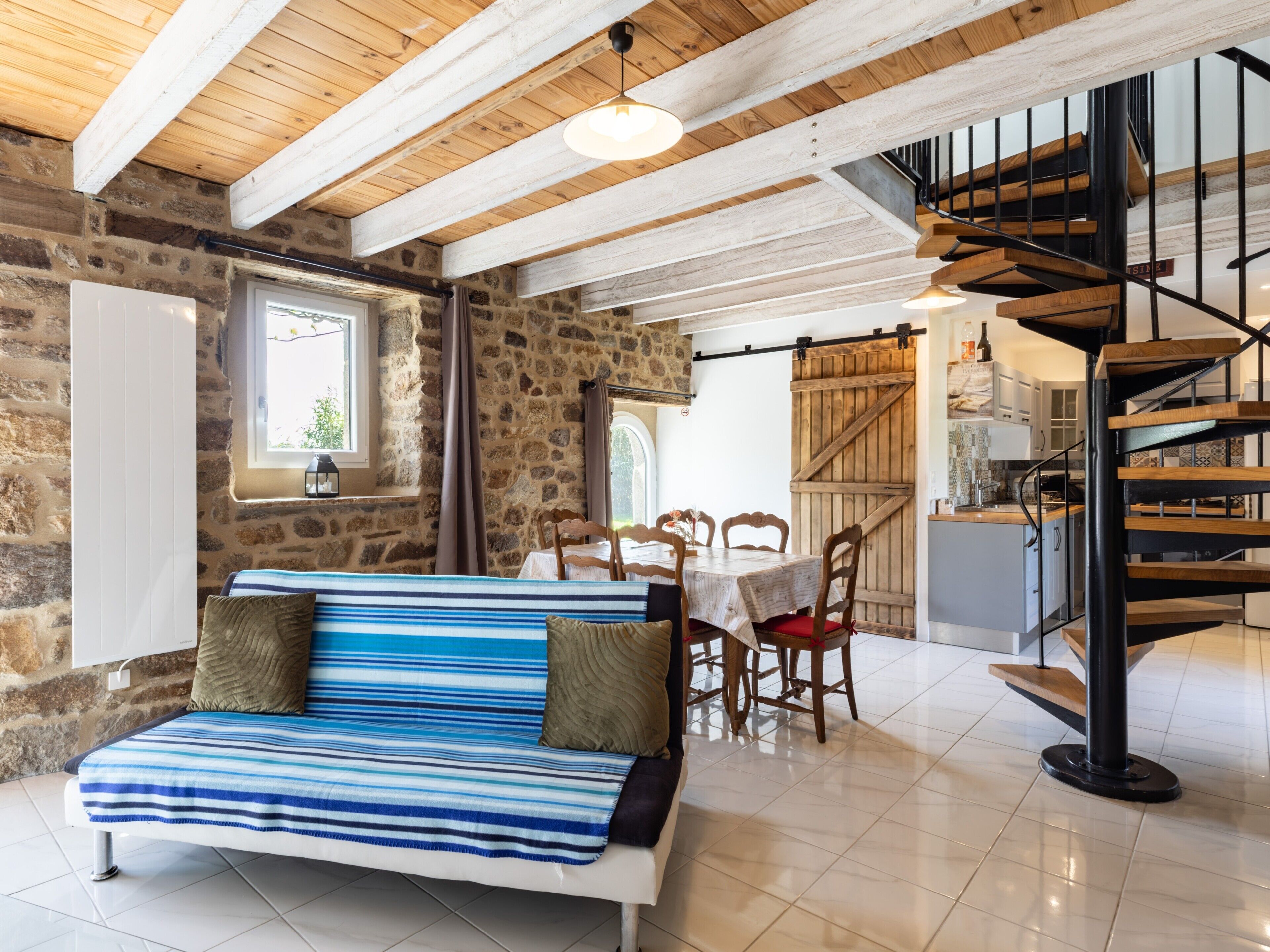 A cozy room with a stone wall, a wooden ceiling, a striped sofa, a dining table with chairs, and a spiral staircase.