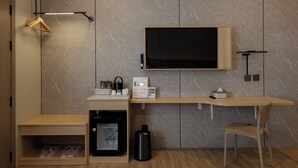 A modern hotel room with a flat-screen TV mounted on the wall, a wooden desk with a chair, a small refrigerator, a shoe rack, and a trash bin. - HippoTel Khaorang Phuket Town (Phuket)