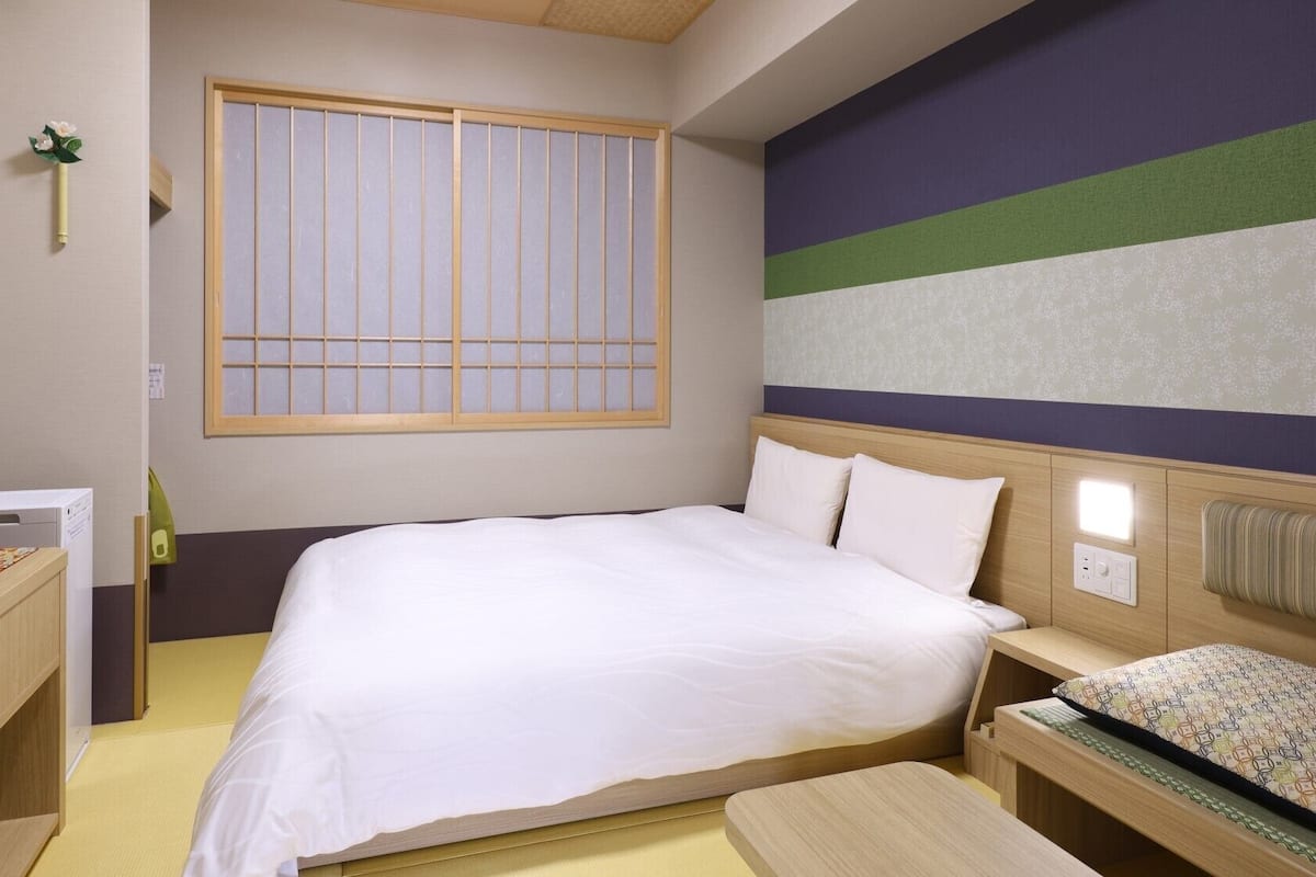 A hotel room with a bed, bedside table, TV, and a window with vertical blinds.
